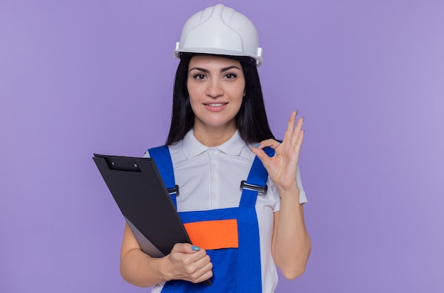 Mujer joven constructor en uniforme de construcción y casco de seguridad con portapapeles y lápiz mirando al frente feliz y positivo sonriendo confiado haciendo bien cartel de pie sobre la pared púrpura