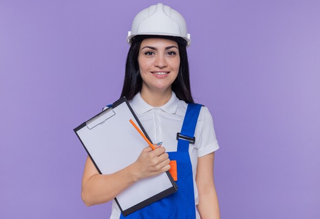 Mujer joven constructor en uniforme de construcción y casco de seguridad con portapapeles y lápiz mirando al frente, feliz y positivo, sonriente, confiado de pie sobre la pared púrpura