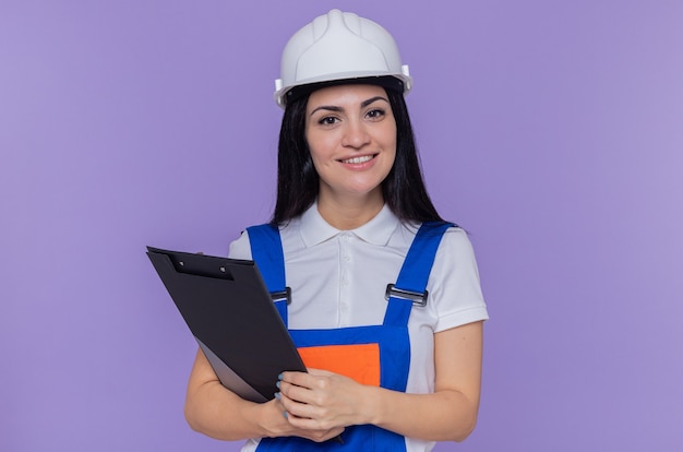 Mujer joven constructor en uniforme de construcción y casco de seguridad con portapapeles y lápiz mirando al frente, feliz y positivo, sonriente, confiado de pie sobre la pared púrpura