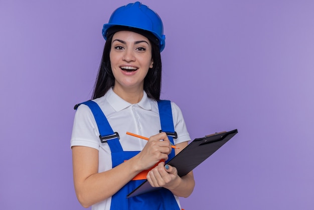 Mujer joven constructor en uniforme de construcción y casco de seguridad con portapapeles y lápiz mirando al frente sonriendo alegremente de pie sobre la pared púrpura