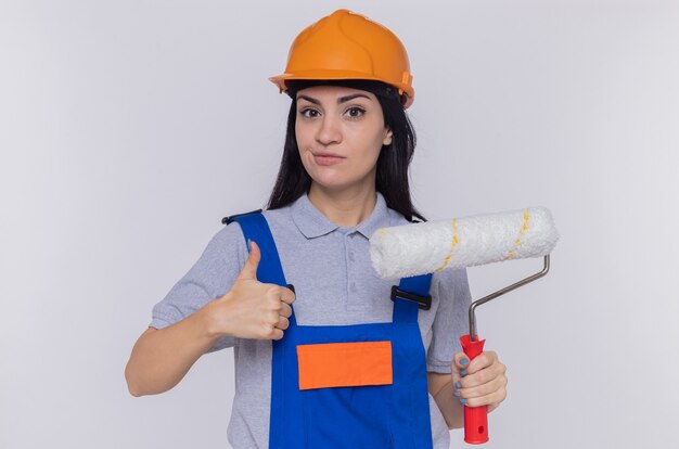 Mujer joven constructor en uniforme de construcción y casco de seguridad con rodillo de pintura mostrando el pulgar hacia arriba sonriendo confiado de pie sobre la pared blanca