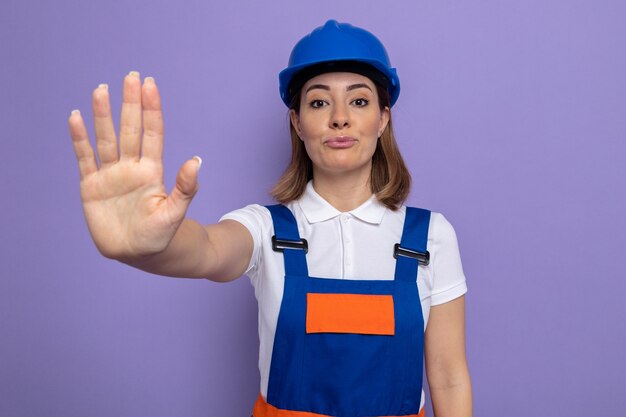 Mujer joven constructor en uniforme de construcción y casco de seguridad con rostro serio haciendo gesto de parada con la mano de pie sobre la pared púrpura