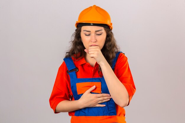 Mujer joven constructor en uniforme de construcción y casco de seguridad sentirse mal y tos sobre la pared blanca aislada