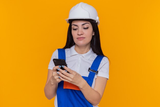 Mujer joven constructor en uniforme de construcción y casco de seguridad con smartphone escribiendo varios mensajes con cara seria de pie sobre la pared naranja