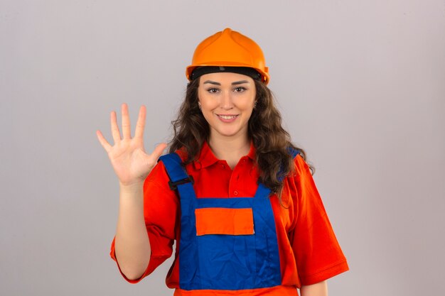 Mujer joven constructor en uniforme de construcción y casco de seguridad sonriendo alegre mostrando y apuntando hacia arriba con los dedos número cinco mirando confiado y feliz sobre la pared blanca aislada