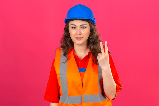 Mujer joven constructor en uniforme de construcción y casco de seguridad sonriendo alegre mostrando y apuntando hacia arriba con los dedos número dos mirando confiado sobre pared rosa aislado