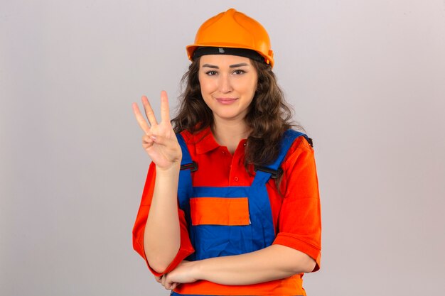 Mujer joven constructor en uniforme de construcción y casco de seguridad sonriendo alegre mostrando y apuntando hacia arriba con los dedos número tres mirando confiado y feliz sobre la pared blanca aislada