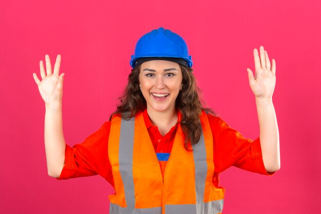 Mujer joven constructor en uniforme de construcción y casco de seguridad sonriendo alegremente con las manos levantadas sobre pared rosa aislado