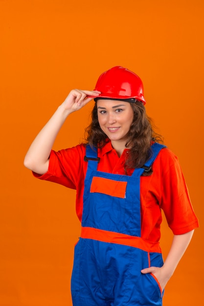 Mujer joven constructor en uniforme de construcción y casco de seguridad sonriendo amable tocando su casco sobre pared naranja aislada