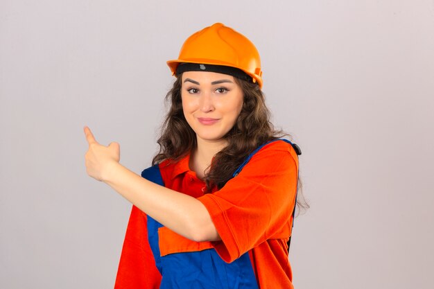 Mujer joven constructor en uniforme de construcción y casco de seguridad sonriendo apuntando con la mano y el dedo hacia un lado sobre la pared blanca aislada