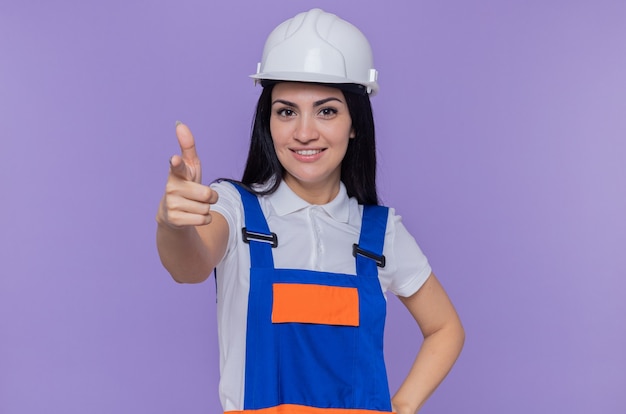 Mujer joven constructor en uniforme de construcción y casco de seguridad sonriendo confiado apuntando con el dedo índice en la parte delantera de pie sobre la pared púrpura