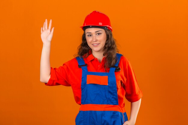 Mujer joven constructor en uniforme de construcción y casco de seguridad sonriendo saludando amigable con mano sobre pared naranja aislada