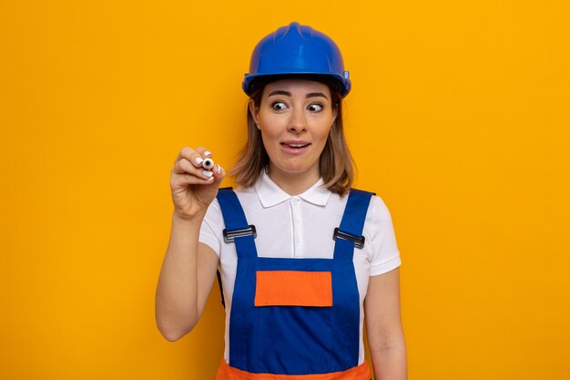 Mujer joven constructor en uniforme de construcción y casco de seguridad con sonrisa astuta en la cara escribiendo con lápiz en el aire de pie sobre la pared naranja