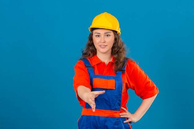 Mujer joven constructor en uniforme de construcción y casco de seguridad con sonrisa ofreciendo mano haciendo gesto de saludo sobre pared azul aislada