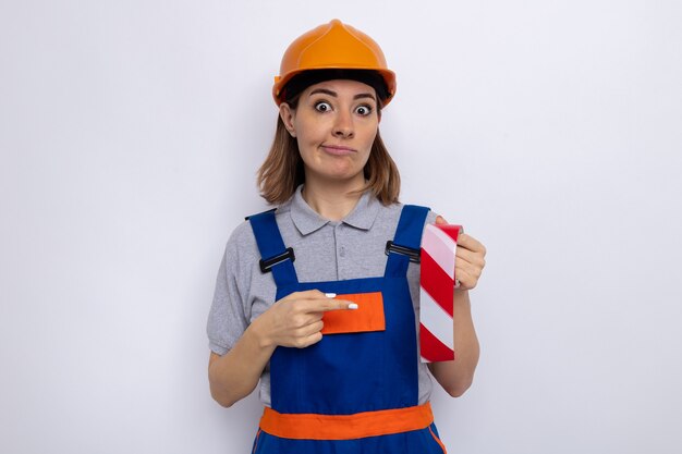 Mujer joven constructor en uniforme de construcción y casco de seguridad sosteniendo cinta adhesiva apuntando con el dedo índice a confundido parado sobre pared blanca