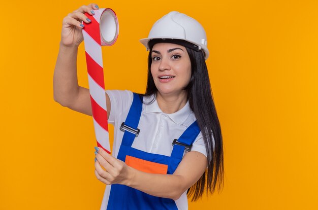 Mujer joven constructor en uniforme de construcción y casco de seguridad sosteniendo cinta adhesiva mirando al frente sonriendo alegremente de pie sobre la pared naranja