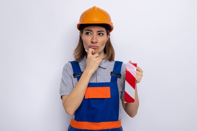 Mujer joven constructor en uniforme de construcción y casco de seguridad sosteniendo cinta adhesiva mirando a un lado con expresión pensativa con la mano en la barbilla de pie sobre la pared blanca