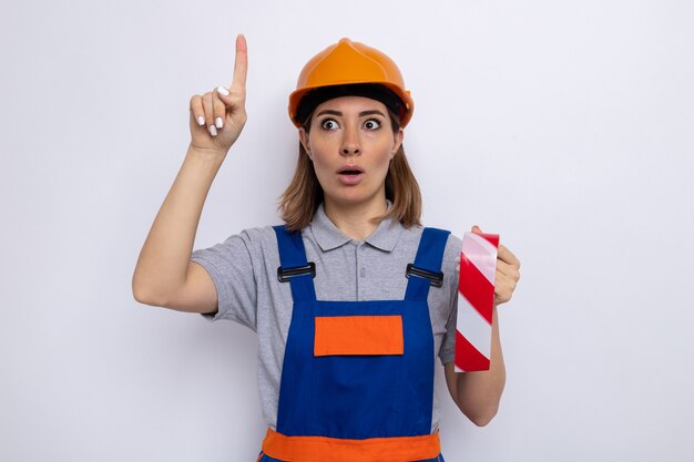 Mujer joven constructor en uniforme de construcción y casco de seguridad sosteniendo cinta adhesiva mirando a un lado sorprendido y preocupado mostrando el dedo índice