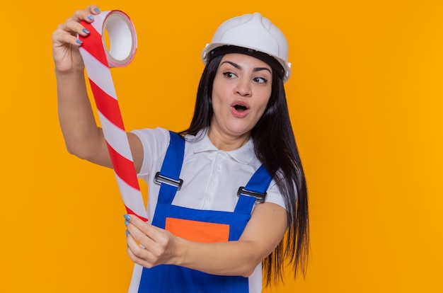 Mujer joven constructor en uniforme de construcción y casco de seguridad sosteniendo cinta adhesiva mirándolo asombrado y sorprendido de pie sobre la pared naranja