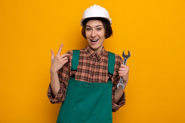 Mujer joven constructor en uniforme de construcción y casco de seguridad sosteniendo una llave feliz y sorprendida mostrando el dedo índice de pie en naranja