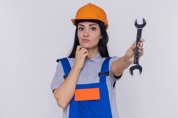 Mujer joven constructor en uniforme de construcción y casco de seguridad sosteniendo una llave mirando al frente desconcertado de pie sobre la pared blanca