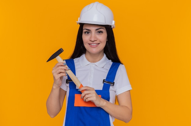 Mujer joven constructor en uniforme de construcción y casco de seguridad sosteniendo un martillo mirando al frente con una sonrisa en la cara de pie sobre la pared naranja