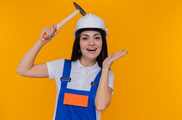 Mujer joven constructor en uniforme de construcción y casco de seguridad sosteniendo un martillo mirando a la cámara