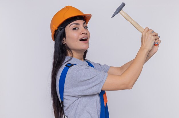 Mujer joven constructor en uniforme de construcción y casco de seguridad sosteniendo un martillo sonriendo confiado mirando al frente de pie sobre la pared blanca