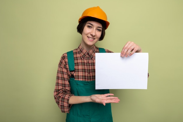 Mujer joven constructor en uniforme de construcción y casco de seguridad sosteniendo la página en blanco que presenta con el brazo de la mano sonriendo alegremente de pie en verde