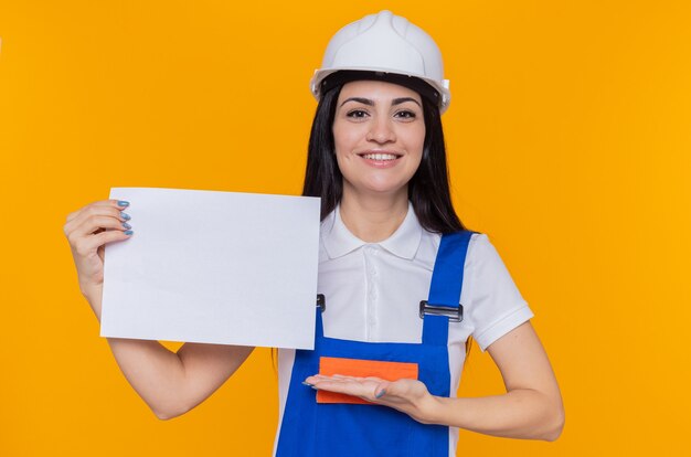 Mujer joven constructor en uniforme de construcción y casco de seguridad sosteniendo la página en blanco que presenta con el brazo de la mano sonriendo confiado de pie sobre la pared naranja