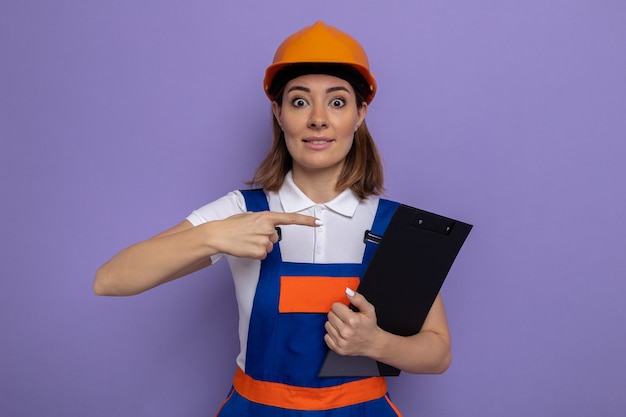 Mujer joven constructor en uniforme de construcción y casco de seguridad sosteniendo el portapapeles apuntando con el dedo índice a él sorprendido y feliz de pie sobre la pared púrpura