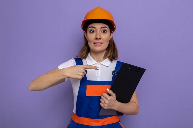 Foto gratuita mujer joven constructor en uniforme de construcción y casco de seguridad sosteniendo el portapapeles apuntando con el dedo índice a él sorprendido y feliz de pie sobre la pared púrpura