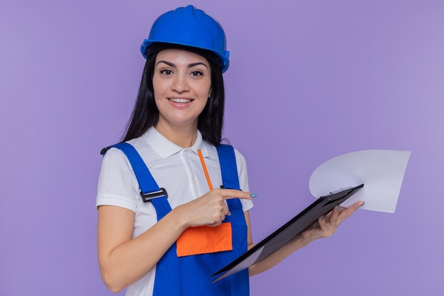 Mujer joven constructor en uniforme de construcción y casco de seguridad sosteniendo portapapeles y lápiz apuntando con el dedo índice en el portapapeles mirando al frente sonriendo alegremente de pie sobre la pared púrpura