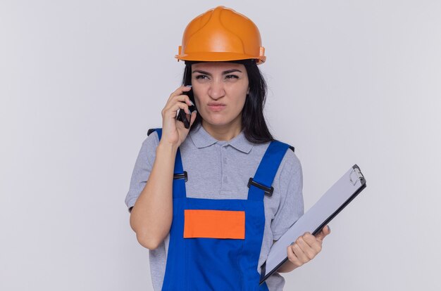 Mujer joven constructor en uniforme de construcción y casco de seguridad sosteniendo portapapeles mirando disgustado mientras habla por teléfono móvil de pie sobre una pared blanca