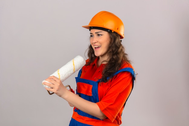 Mujer joven constructor en uniforme de construcción y casco de seguridad usando rodillo de pintura como micrófono cantando canción diversión en el concepto de trabajo sobre pared blanca aislada