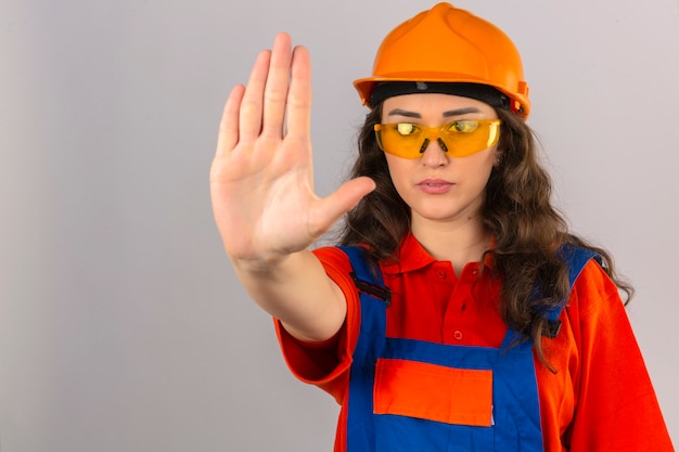 Mujer joven constructor en uniforme de construcción gafas amarillas y casco de seguridad haciendo parada cantar con la palma de la mano expresión de advertencia sobre la pared blanca aislada
