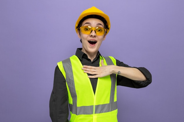 Mujer joven constructora en chaleco de construcción y casco de seguridad con gafas amarillas de seguridad feliz y sorprendido sosteniendo la mano en su pecho de pie sobre la pared azul