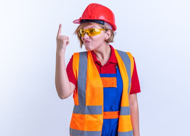 Mujer joven constructora codiciosa en uniforme con gafas mostrando uno aislado sobre fondo blanco.