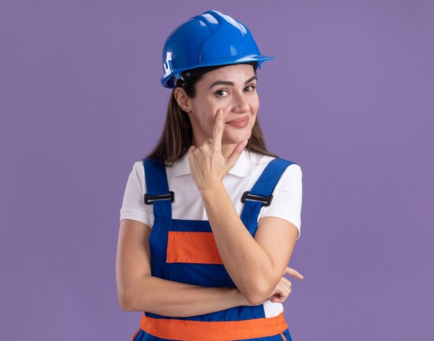 Mujer joven constructora complacida en uniforme bajó el párpado aislado en la pared púrpura