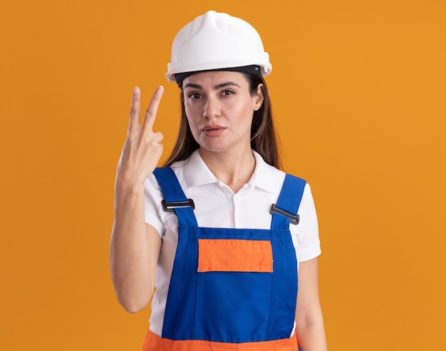 Mujer joven constructora complacida en uniforme mostrando gesto de paz aislado en pared naranja