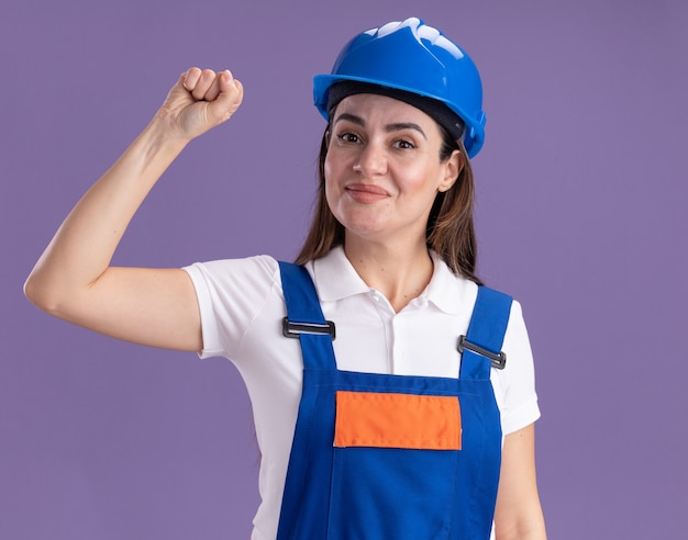 Mujer joven constructora complacida en uniforme mostrando gesto sí aislado en la pared púrpura