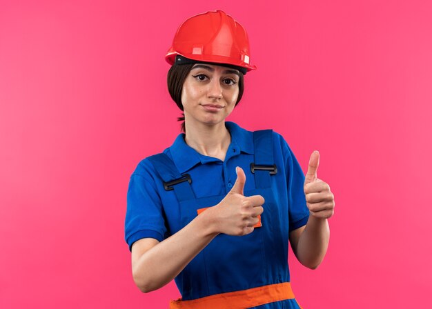 Mujer joven constructora complacida en uniforme mostrando los pulgares para arriba aislado en la pared rosa