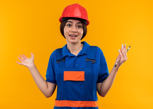 Mujer joven constructora complacida en uniforme sosteniendo una llave de boca extendiendo las manos aisladas en la pared amarilla