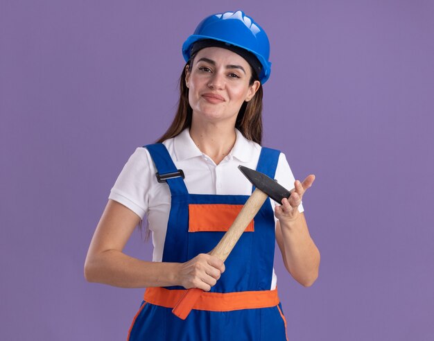 Mujer joven constructora complacida en uniforme sosteniendo un martillo aislado en la pared púrpura