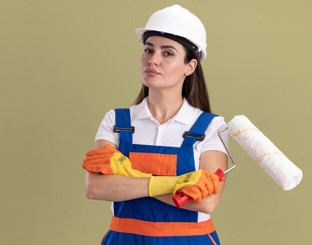 Mujer joven constructora confiada en uniforme y guantes sosteniendo un cepillo de rodillo y cruzando las manos aisladas en la pared verde oliva