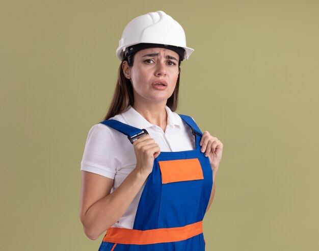 Mujer joven constructora disgustada en uniforme agarró uniforme aislado en la pared verde oliva