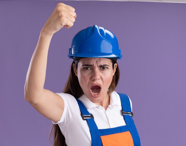Mujer joven constructora emocionada en uniforme levantando el puño aislado en la pared púrpura