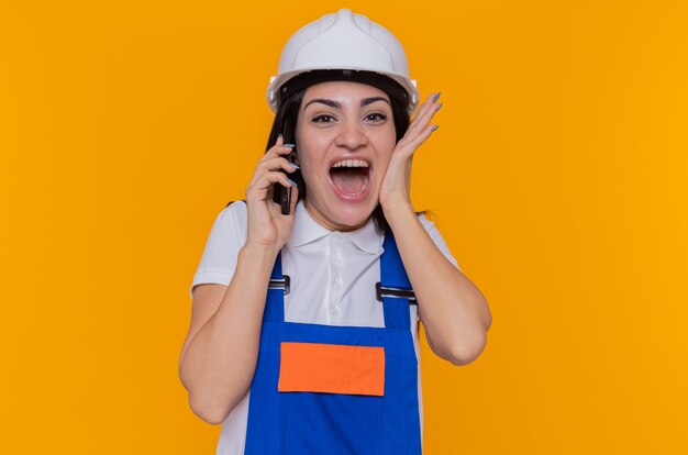 Mujer joven constructora feliz y emocionada loca en uniforme de construcción y casco de seguridad hablando por teléfono móvil de pie sobre la pared naranja