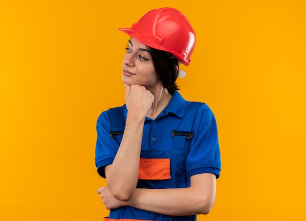 Mujer joven constructora de lado mirando impresionado en uniforme poniendo la mano debajo de la barbilla aislada en la pared amarilla