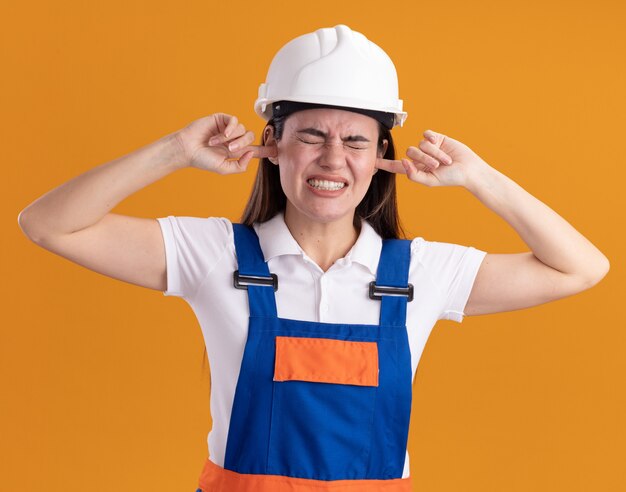 Mujer joven constructora molesta en orejas cubiertas uniforme aislado en pared naranja